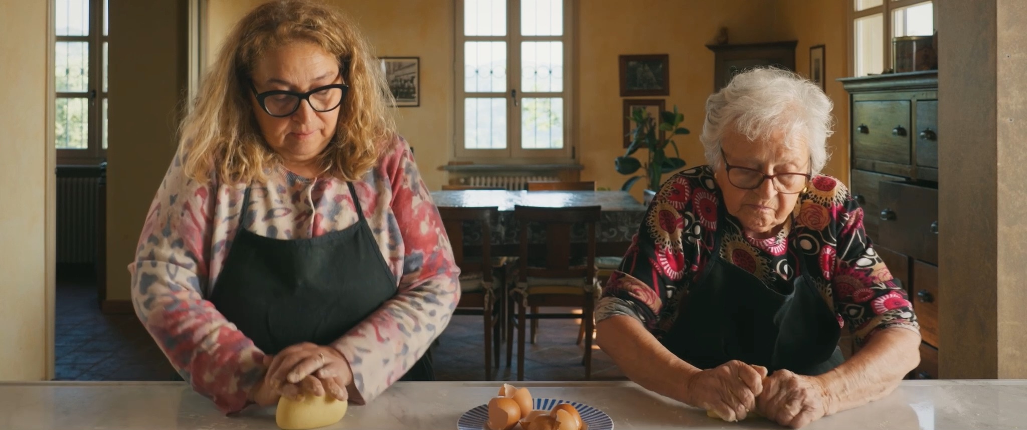 Susanna and grandmother Leila knead dough side by side at the kitchen counter, a plate of cracked eggshells between them.